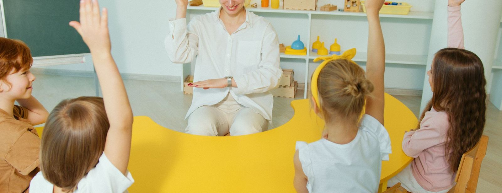 Erzieherin sitzt im Morgenkreis, Kinder melden sich mit erhobenen Armen